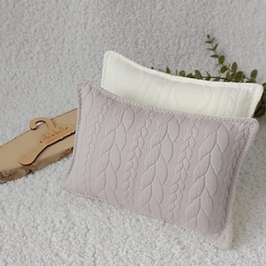 Op de afbeelding: Twee decoratieve kussens met een kabelbreipatroon. Het bovenste kussen is wit en het onderste kussen is lichtpaars.