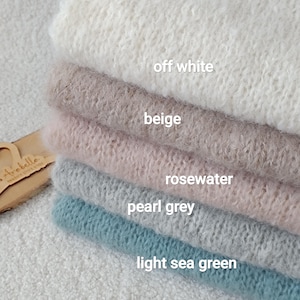 Peut inclure: Une pile de cinq textiles en tricot duveteux pliés dans les tons blanc cassé, beige, rose poudré, gris perle et vert d'eau clair. Chaque couche est étiquetée avec son nom de couleur en blanc. Une petite étiquette en bois porte le nom Arabella.