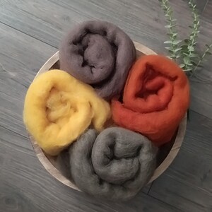 Puede incluir: Un cuenco de madera lleno de cinco rollos de lana de colores. La lana es en tonos de amarillo, naranja, gris y morado. El cuenco está sobre una superficie de madera gris. Una ramita de vegetación está en el fondo.
