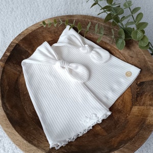 Peut inclure: Deux bandeaux en tissu blanc côtelé avec un nœud central. Chaque bandeau est fabriqué dans une matière souple et extensible, orné d'un petit bouton en bois circulaire sur le côté et d'une délicate bordure en dentelle blanche sur le bord.