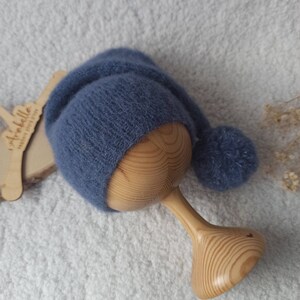 Puede incluir: Un gorro de bebé de punto azul suave con una parte superior larga y puntiaguda y un pompón a juego. El gorro se exhibe en un soporte de madera. El gorro está hecho de un material difuso.