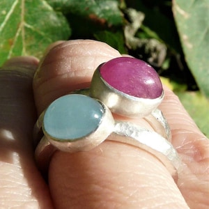 May include: Two silver rings with gemstones. The ring on the left has a light blue aquamarine stone. The ring on the right has a pink ruby stone.