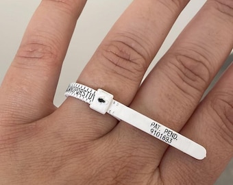 Pode incluir: Um medidor de tamanho de anel de plástico branco com marcas pretas para o tamanho. O medidor está em um dedo e o texto "PAT. PEND. 9101693" está impresso no medidor.