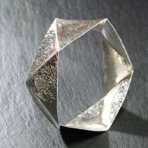 Puede incluir: Un brazalete geométrico de plata con acabado martillado. El brazalete está abierto y tiene forma hexagonal.