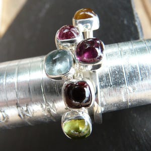 Op de afbeelding: Een zilveren ring met zes gekleurde edelstenen. De edelstenen zijn in een cluster gerangschikt en zijn gezet in zilver. De ring ligt op een zilveren meetlint.