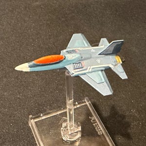 Puede incluir: Un modelo de avión en miniatura en tonos azules, naranjas y amarillos, montado sobre un soporte transparente. El avión de juguete tiene un diseño elegante con marcas detalladas y es probablemente un objeto de colección o una pieza de juego.