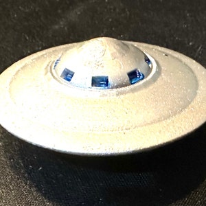 Może przedstawiać: Srebrna zabawka w kształcie latającego spodka z niebieskimi detalami wokół krawędzi. Zabawka jest wykonana z metalu i ma gładkie, błyszczące wykończenie.