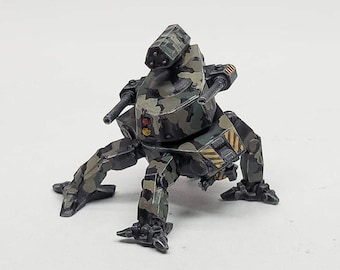 May include: A miniature military mech toy with a camouflage pattern in shades of green, brown, and gray. The toy has a central body with a turret and two side-mounted cannons. The legs are articulated, and the overall design suggests a futuristic war machine.