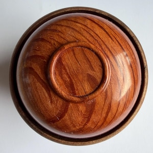 May include: A wooden bowl and lid, crafted from reddish-brown wood. The bowl and lid display a prominent wood grain pattern. The rim of the bowl is a darker shade of brown. The lid features a circular indentation in the centre.