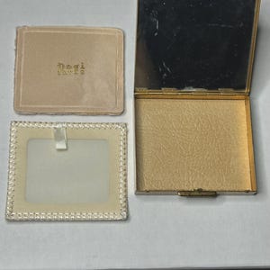 May include: An open, gold-toned compact with a mirror and a textured interior, next to a small, beige fabric case with the word "Paris" embroidered on it. A small, rectangular frame with a white insert and a ribbon is also present.