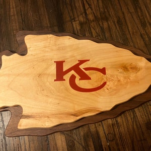 May include: A wooden cutting board shaped like an arrowhead with a red "KC" logo in the center. The board is made of two types of wood, a lighter wood for the arrowhead and a darker wood for the border.