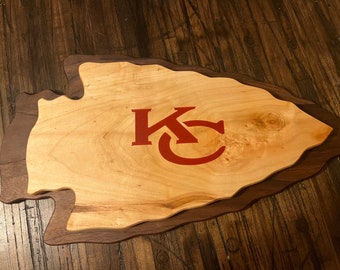 Op de afbeelding: Een houten snijplank in de vorm van een pijlpunt met een rood "KC" logo in het midden. De plank is gemaakt van twee soorten hout, een lichter hout voor de pijlpunt en een donkerder hout voor de rand.