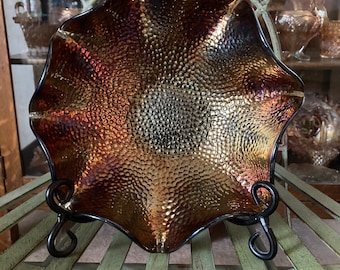 May include: A large, iridescent glass bowl with a hammered texture. The bowl is a deep brown color with hints of gold and purple. It is sitting on a black metal stand.