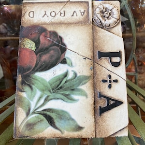 May include: A decorative tile featuring a detailed floral design with a large, dark red flower and green leaves. The tile has a distressed, aged appearance with the letters "A" and "P" visible, along with the text "AV ROY DA".