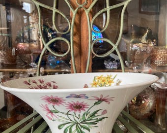 May include: White ceramic bowl with a flared design, adorned with pink, yellow, and purple floral illustrations and butterflies. The bowl is labeled "Gazania Ringens, Treasure Flower". The bowl is sitting on a green metal stand.