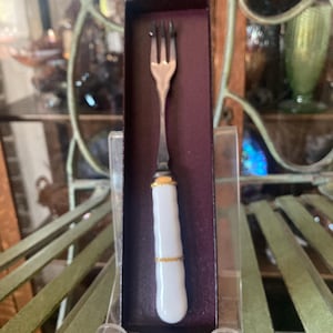 May include: A three-tined silver-toned metal fork with a white ceramic handle featuring vertical ridges and two thin gold-colored bands. The fork is presented inside a narrow, dark-colored rectangular box standing upright.