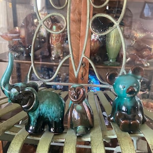 May include: Three ceramic animal figurines: an elephant, an owl, and a mouse. The elephant and mouse are teal and black, while the owl is brown and black. They are displayed on a green metal shelf, with a glass cabinet in the background.