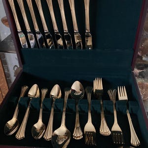 May include: A gold-toned flatware set housed in a dark wooden storage case with a dark green velvet interior. The set includes eight knives arranged vertically in the lid, and a collection of spoons and forks organized in the base.