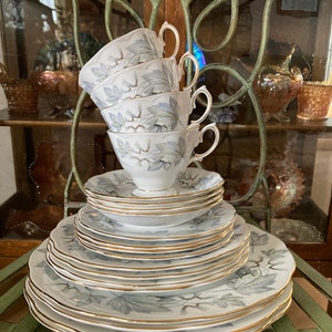 May include: A set of white china dinnerware with a grey and silver leaf pattern and gold trim. The set includes plates, saucers, and teacups.
