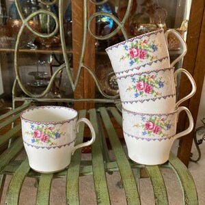 Peut inclure: Quatre tasses à thé blanches avec des motifs floraux roses et bleus empilées sur une chaise en métal verte. Les tasses ont un motif floral délicat et un bord doré.