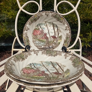 May include: Two white ceramic bowls featuring a brown, green, and pink transferware landscape scene of a house and trees. The bowls have a scalloped, swirled rim with a detailed leaf border pattern. The design is repeated on both pieces.