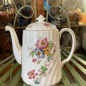 May include: White ceramic teapot with a floral design in shades of pink, yellow, blue, and green. The teapot has a gold trim around the lid and spout.