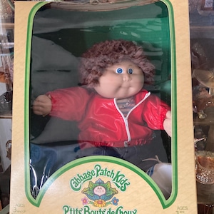 May include: A Cabbage Patch Kids doll in its original packaging. The doll has brown curly hair, blue eyes, and is wearing a red jacket and blue jeans. The box features the words "Cabbage Patch Kids" and "Ptits Bouts de Choux".