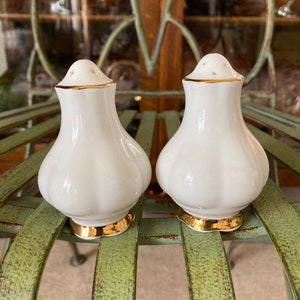 May include: A pair of white ceramic salt and pepper shakers with gold trim. The shakers have a ribbed design and are sitting on a green metal surface.