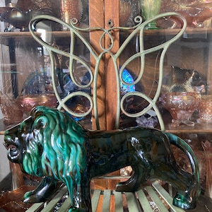 May include: A ceramic lion statue with a green and black glaze. The lion is in a walking pose. The statue is displayed on a green slatted surface, with a decorative metal butterfly in the background. The lion's mane has a textured appearance.
