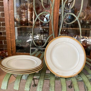 May include: Four white dinner plates with a gold trim stacked on a green metal bench. The plates are made of fine china and have a delicate design.