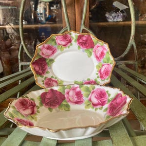 May include: Two white ceramic dishes featuring a rose pattern. The roses are pink and red with green foliage. The edges of the dishes are scalloped and trimmed with gold. One is oval, the other a shallow bowl, ideal for serving.