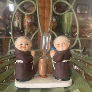 May include: A vintage hourglass figurine featuring two smiling monk figures in brown robes flanking a central glass hourglass. The base is white, and the figures have painted facial features.