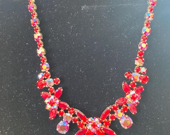 May include: A vintage red rhinestone necklace with a gold-tone metal chain. The necklace features a floral design with various sizes of red and iridescent rhinestones. The necklace is displayed on a black surface.