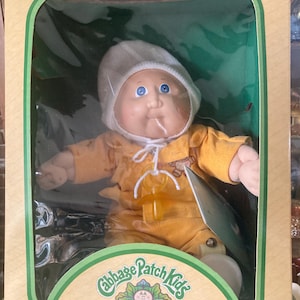 May include: A Cabbage Patch Kids doll with blue eyes and a white knit cap, wearing a yellow outfit with a small graphic. The doll has a yellow pacifier around its neck and is enclosed in a cardboard box with green trim and the text Cabbage Patch Kids.