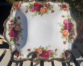 May include: A square porcelain dish featuring a white center and a border decorated with clusters of red and yellow roses with green leaves. The edges of the dish are scalloped and finished with a decorative gold trim along the entire perimeter.