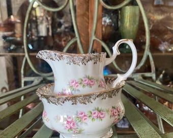 May include: A white porcelain creamer and sugar bowl set featuring a delicate pink rose floral pattern and ornate gold trim along the scalloped rims. The creamer has a curved handle and a pouring spout, while the sugar bowl is round.