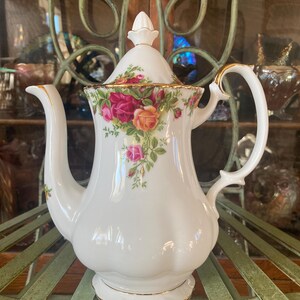 May include: White ceramic teapot with a floral rose design in pink, red, and yellow. The teapot has a gold trim and a curved handle. The lid is topped with a small finial.