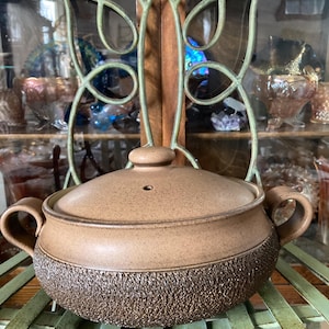 May include: A brown ceramic casserole dish with a lid and two handles. The bottom half of the dish has a textured, rough surface. The dish is sitting on a green metal decorative stand.