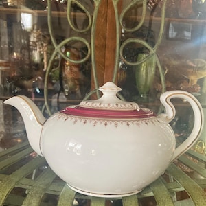 Peut inclure: Théière en porcelaine blanche avec un bord et des accents bordeaux et dorés. La théière a un bec et une anse incurvés, et un couvercle assorti avec un bouton doré. La théière est sur une étagère en métal vert.