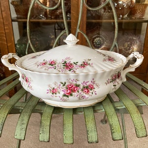 May include: White porcelain serving bowl with a lid, featuring a floral pattern of pink roses and green leaves. The bowl has a gold trim around the rim.