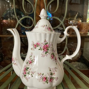 May include: A white porcelain teapot with a floral pattern of pink roses and green leaves. The teapot has a gold trim around the lid and base.