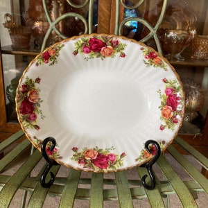 May include: A white porcelain plate with a gold rim and a floral design featuring red, pink, and yellow roses. The plate is on a green metal stand.