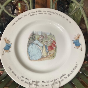 May include: White ceramic plate featuring a scene from a children's story with rabbits. The plate has a central illustration of rabbits and text around the rim. Two rabbits in blue jackets are on the sides. The plate is on a decorative metal stand.