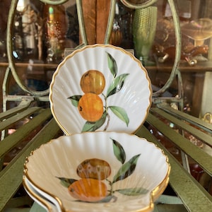 May include: Two white, scalloped-edge dishes with a gold trim. Each dish features a painted design of oranges and green leaves. The dishes are arranged on a green metal surface, showcasing a vintage aesthetic.