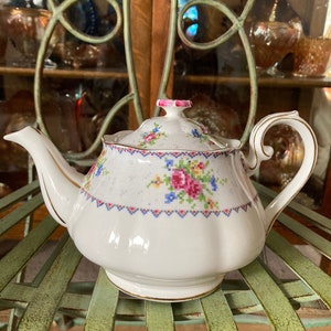 Peut inclure: Théière blanche avec un motif floral et une bordure dorée. La théière a un couvercle rose et un motif floral délicat avec des fleurs roses, bleues et jaunes. La théière est posée sur une chaise en osier verte.