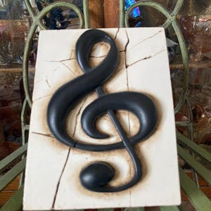 May include: A black treble clef sculpture on a rectangular, off-white tile with a cracked surface. The musical symbol is raised and has a glossy finish. The tile has a brown border. The background is a decorative metal structure.