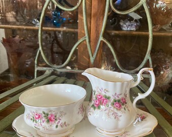 May include: A white porcelain sugar bowl and creamer set with a floral pattern featuring pink roses and green leaves. The set is on a matching oval tray with a gold trim.