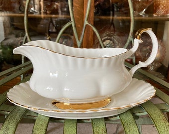 May include: White porcelain gravy boat and matching saucer with gold trim. The gravy boat has a curved handle and a scalloped rim. The saucer has a gold accent in the center. The set is placed on a green metal surface.
