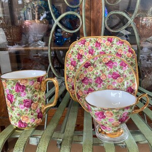 May include: A set of vintage teacups and saucers with a floral pattern. The cups and saucers are decorated with pink and yellow roses on a green background. The set includes a teacup, saucer, and a mug.