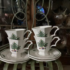 May include: Four white teacups and saucers with a Christmas tree design. The cups have a unique handle shape and sit on matching saucers with a green border. The teacups and saucers are arranged on a green metal surface.